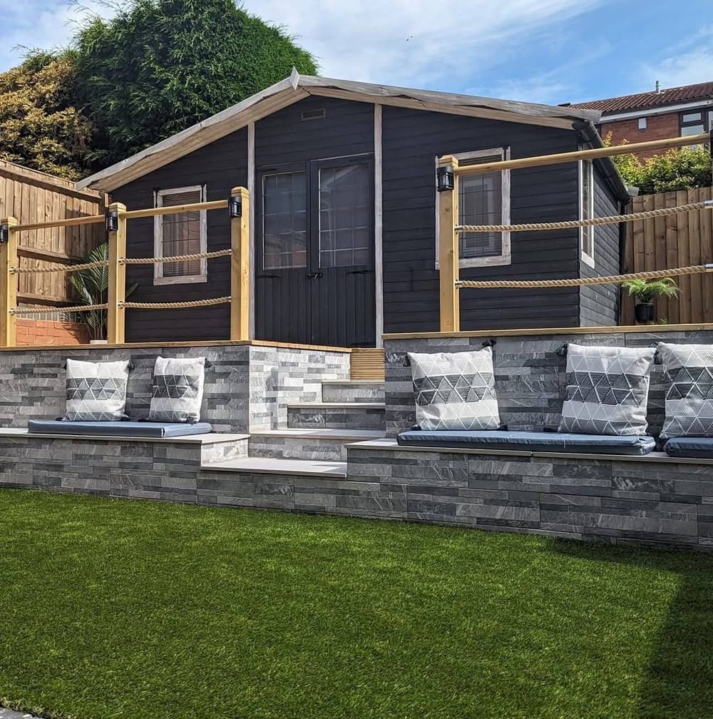 Dark timber garden room with a rope and post balustrade above a grey stone-clad seating wall with cushions, on an artificial lawn