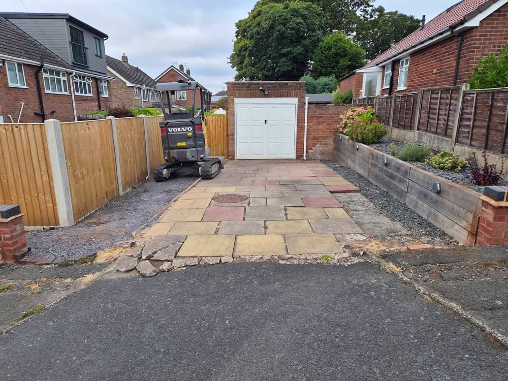 Old paving slabs broken up and a mini excavator working on a driveway in front of a garage, beside a sleeper-edged raised bed