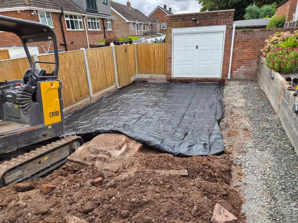Black membrane laid over the dug-out driveway base in front of a garage, with a mini excavator in the foreground