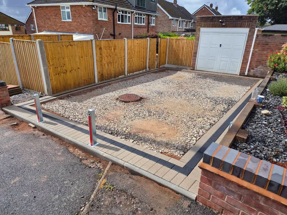Stone base laid inside block edging with a red and white bollard and a blue-brick-topped pier, in front of a garage and timber fence