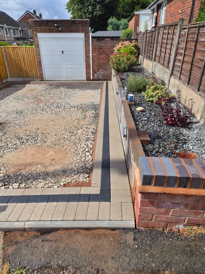 Stone base running up to a garage beside block edging, with a sleeper raised bed of plants and a blue-brick-topped brick pier
