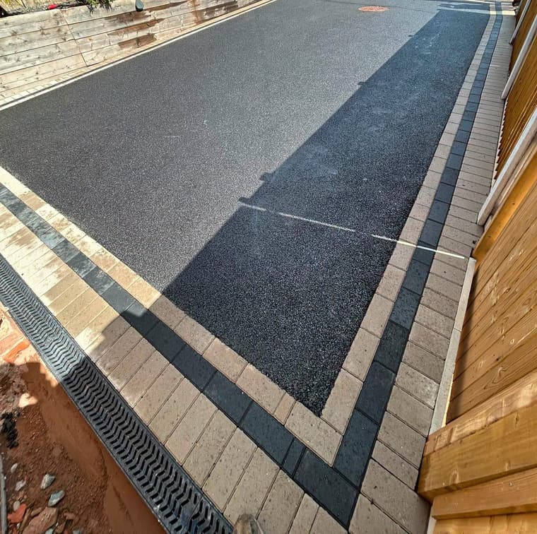 Close view of newly laid black tarmac with buff and charcoal block edging and a drainage channel, beside a timber fence