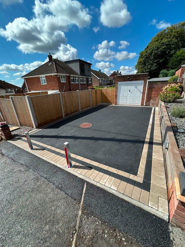 Finished black tarmac driveway with block edging and a bollard, seen from the street under a blue sky with clouds