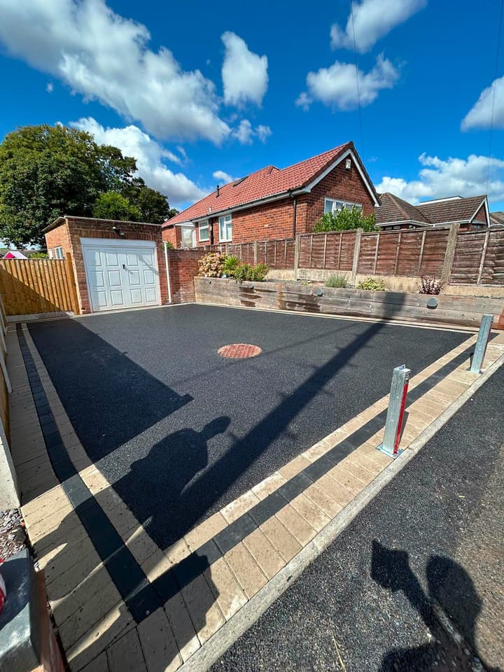 Finished tarmac driveway with block edging, bollards and a manhole cover in front of a garage, under blue sky