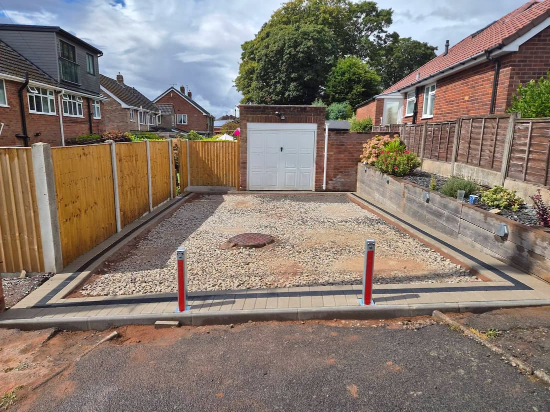 Gravel stone base laid for a driveway in front of a white garage door, with block edging, two red and white bollards, a manhole cover and a timber fence