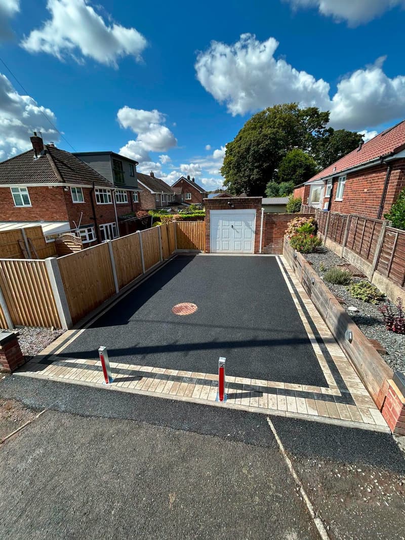 Finished black tarmac driveway with buff block edging, two red and white bollards and an orange manhole cover in front of a garage, under blue sky