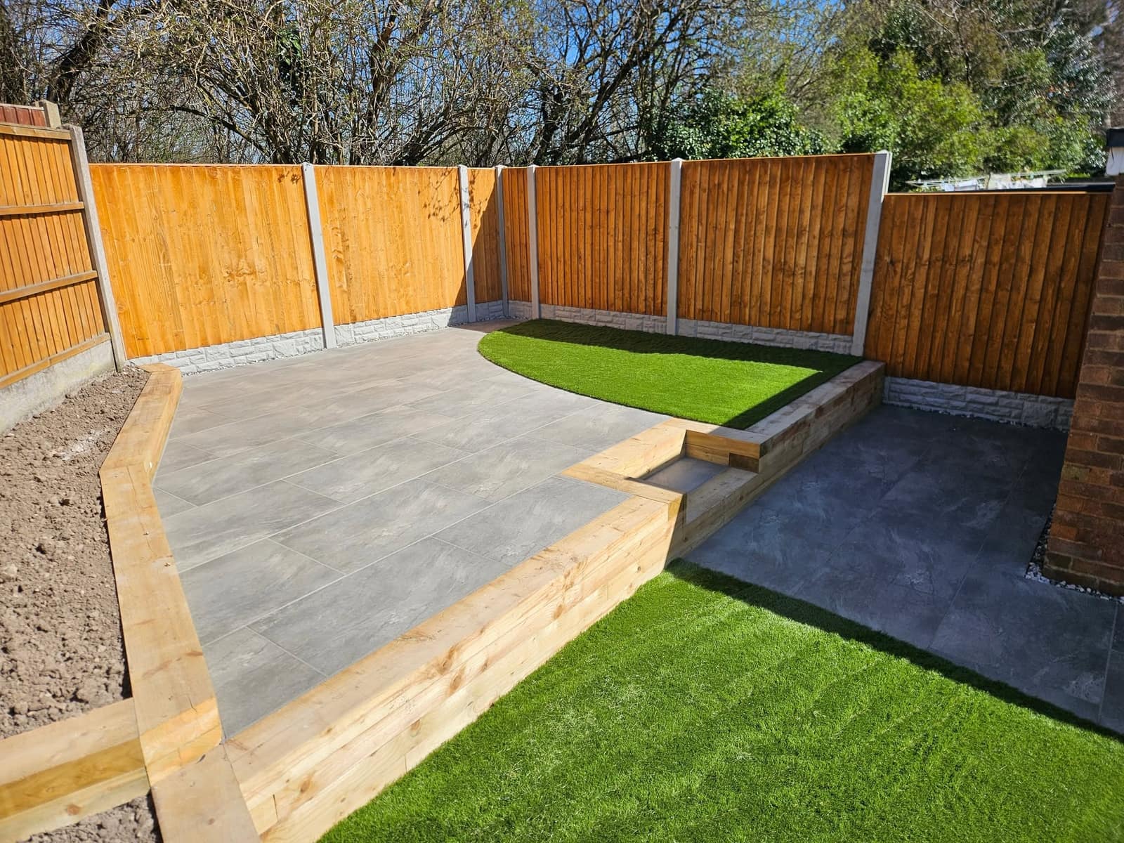 Grey porcelain patio edged with timber sleepers, a raised bed of artificial lawn and honey-coloured fence panels on concrete posts