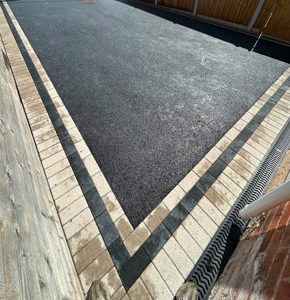 Close view of black tarmac edged with buff and charcoal blocks, with a timber sleeper wall on one side and a drainage channel on the other